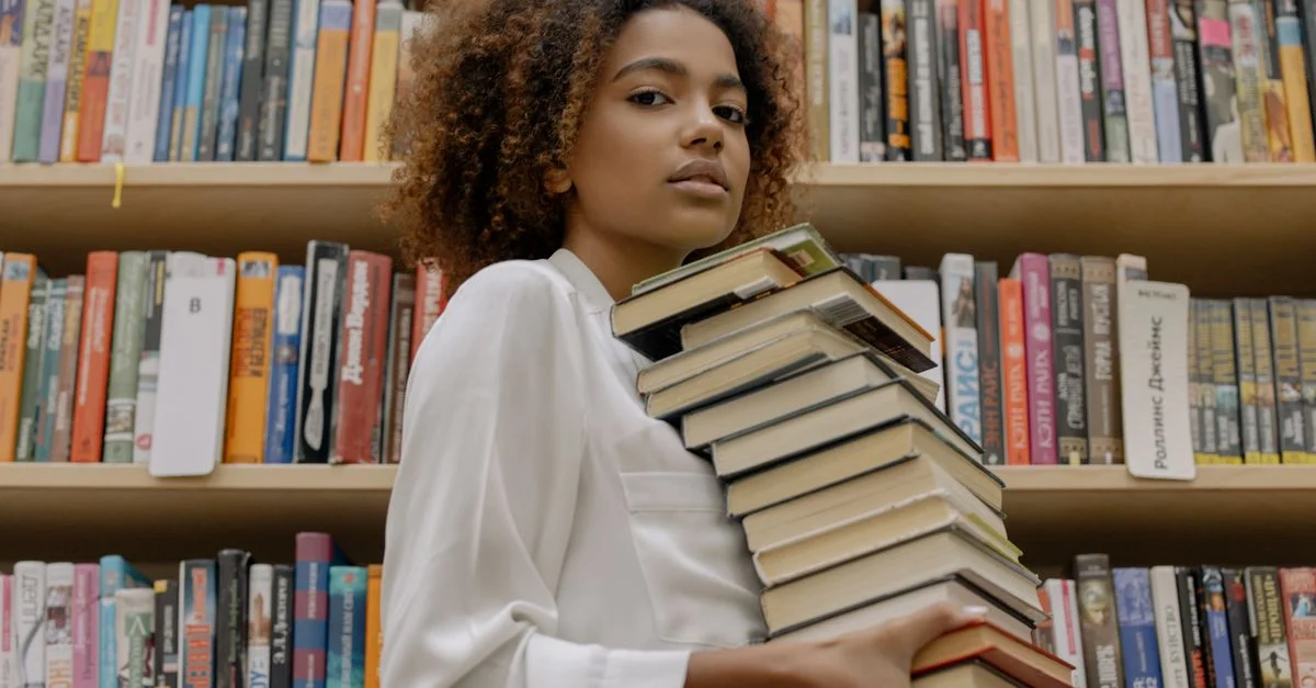 Jeune femme confiante tient une pile de livres dans un cadre de bibliothèque.