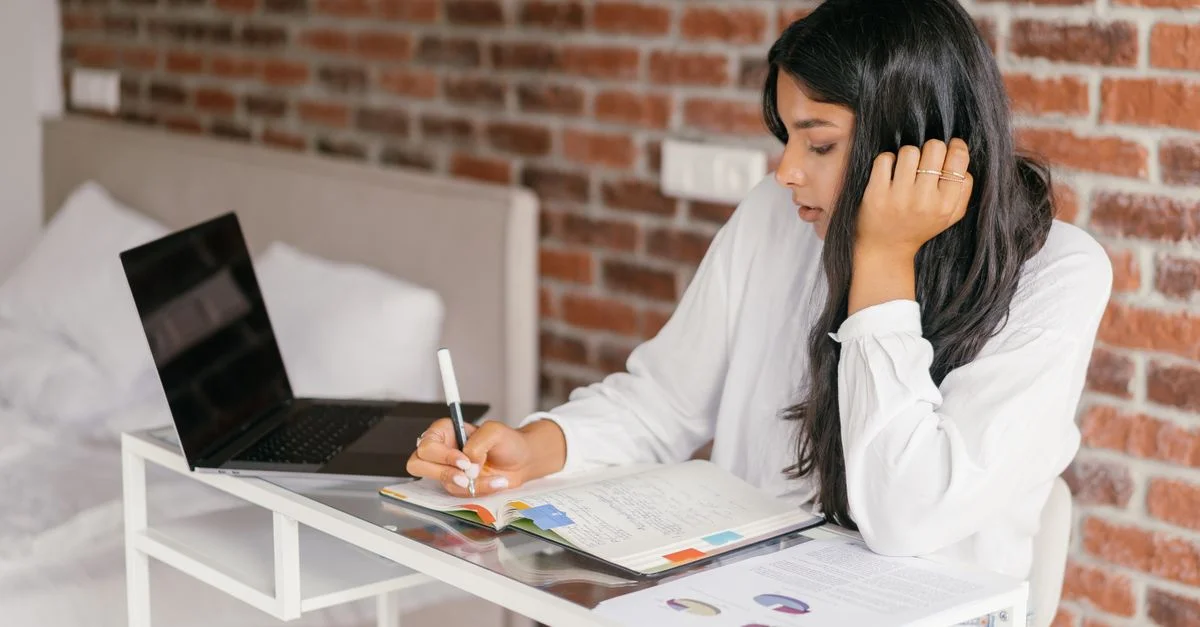 Une jeune femme étudie à l'intérieur avec un ordinateur portable, prenant des notes dans un carnet sur un bureau.