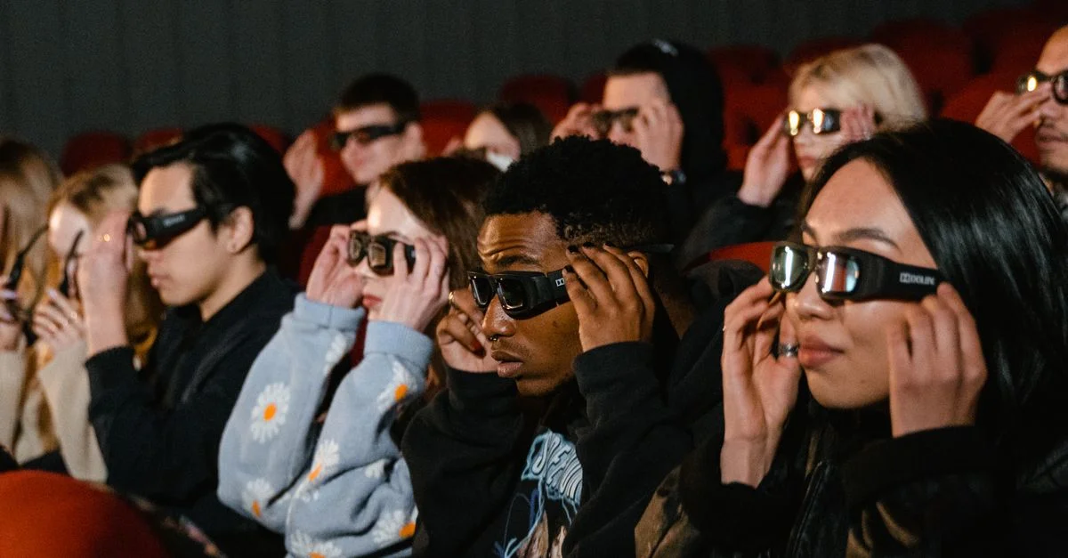Un groupe diversifié de personnes portant des lunettes 3D regardant un film dans un cinéma avec des sièges rouges.
