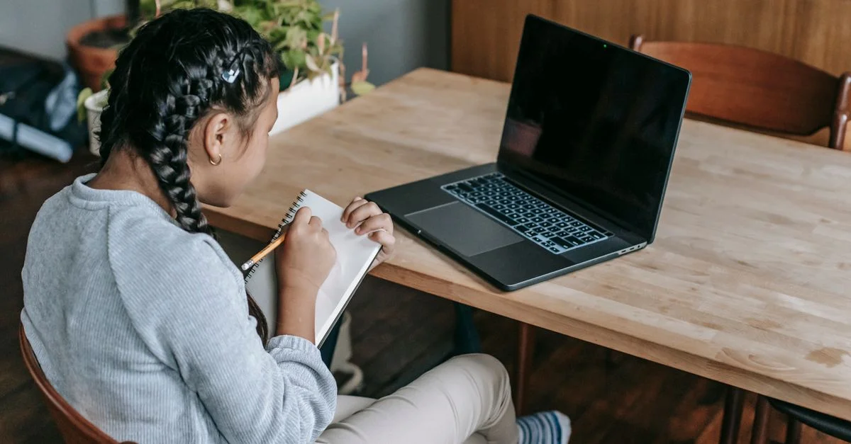 Jeune fille concentrée sur ses devoirs avec un ordinateur portable et un carnet dans une pièce confortable.