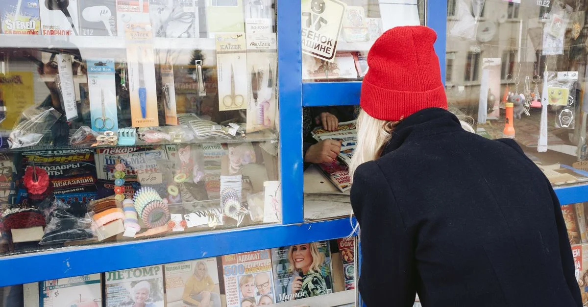 Une femme portant un chapeau rouge feuillette des magazines dans un kiosque extérieur présentant divers articles.